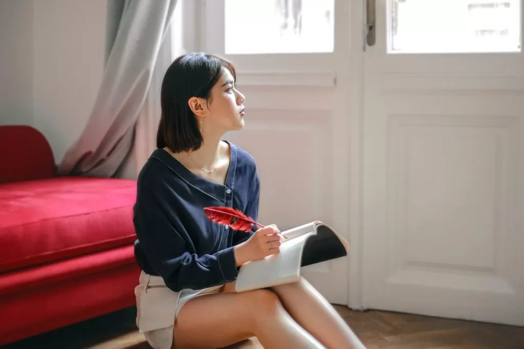 Woman sits at foot of red bed writing in journal with feather quill