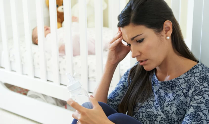 Mother sitting down beside baby's crib, looking upset, symbolizing postpartum depression