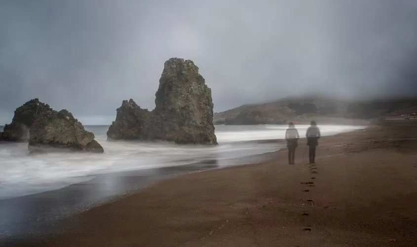 Two faint, ghost-like figures walking along a foggy beach with large rock formations