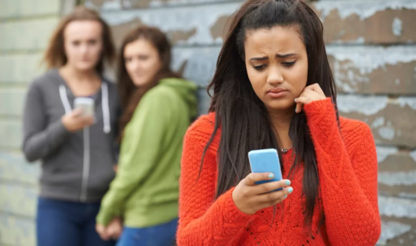 Teen girl on phone, with two teen girls behind on phone, symbolizing online bullying 