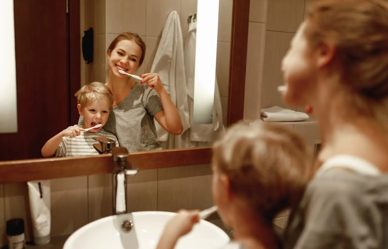 Mother and young child brushing their teeth together in a bathroom while smiling at their reflection in the mirror.
