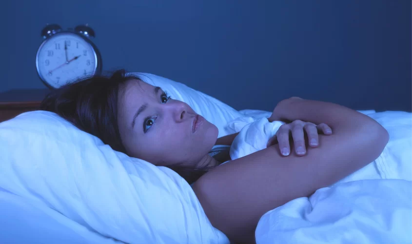 Woman laying in bed with eyes open, arms crossed, alarm clock behind her, symbolizing insomnia
