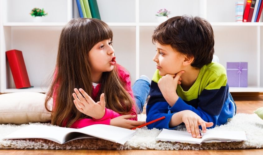 Children laying on ground with books open communicating with one another