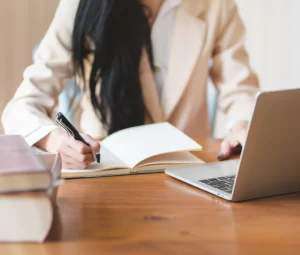 Person writing notes in a notebook beside a laptop.