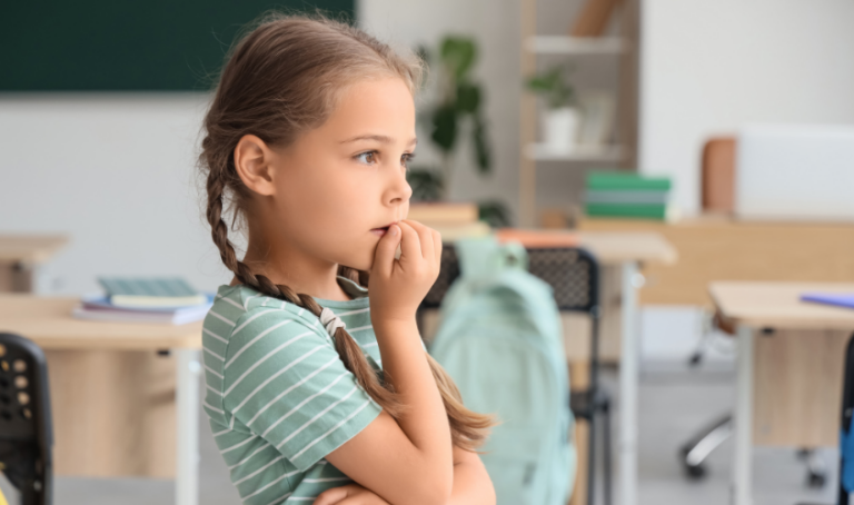 Young girl standing in a classroom biting her nails anxiously.