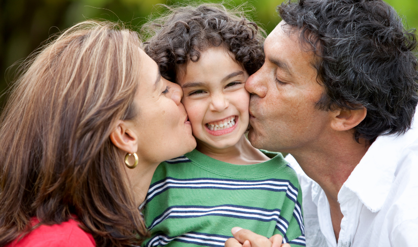 A mother and father kiss their child on the cheek at the same time