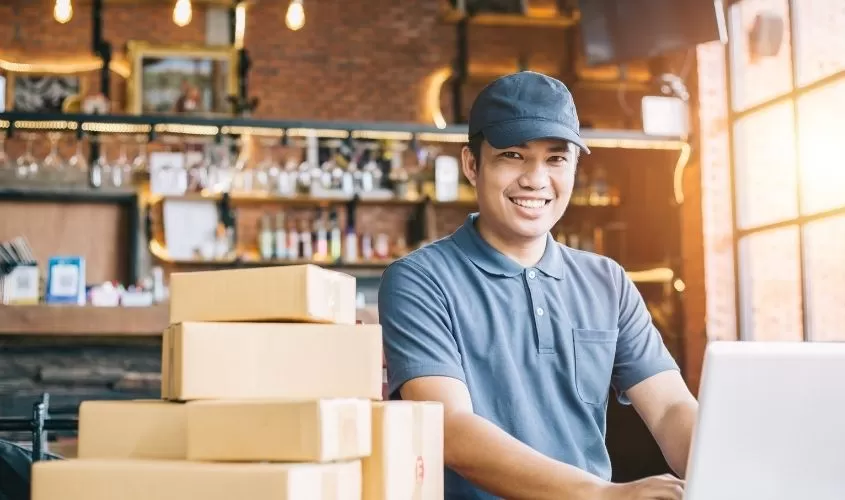 Man smiling, wearing a cap, working on a laptop surrounded by shipping boxes