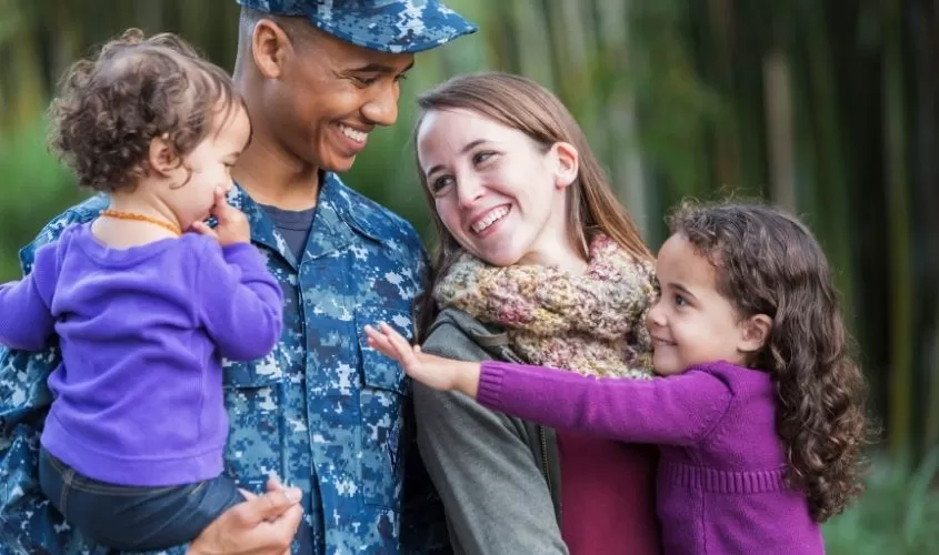 Smiling military family embracing outdoors, with a uniformed service member, partner, and two children