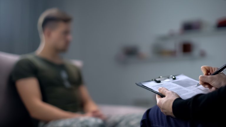 Therapist writing notes with a male client in the background