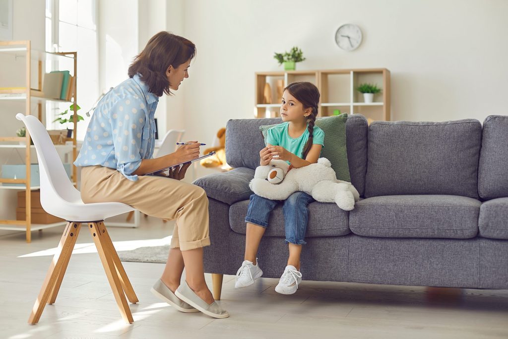 A therapist sits on a chair while attentively listening to a young girl holding a stuffed toy, seated on a couch during a counseling session in a bright and cozy room.