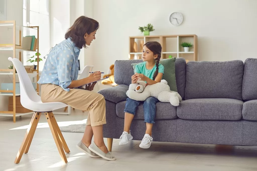 A therapist sits on a chair while attentively listening to a young girl holding a stuffed toy, seated on a couch during a counseling session in a bright and cozy room.