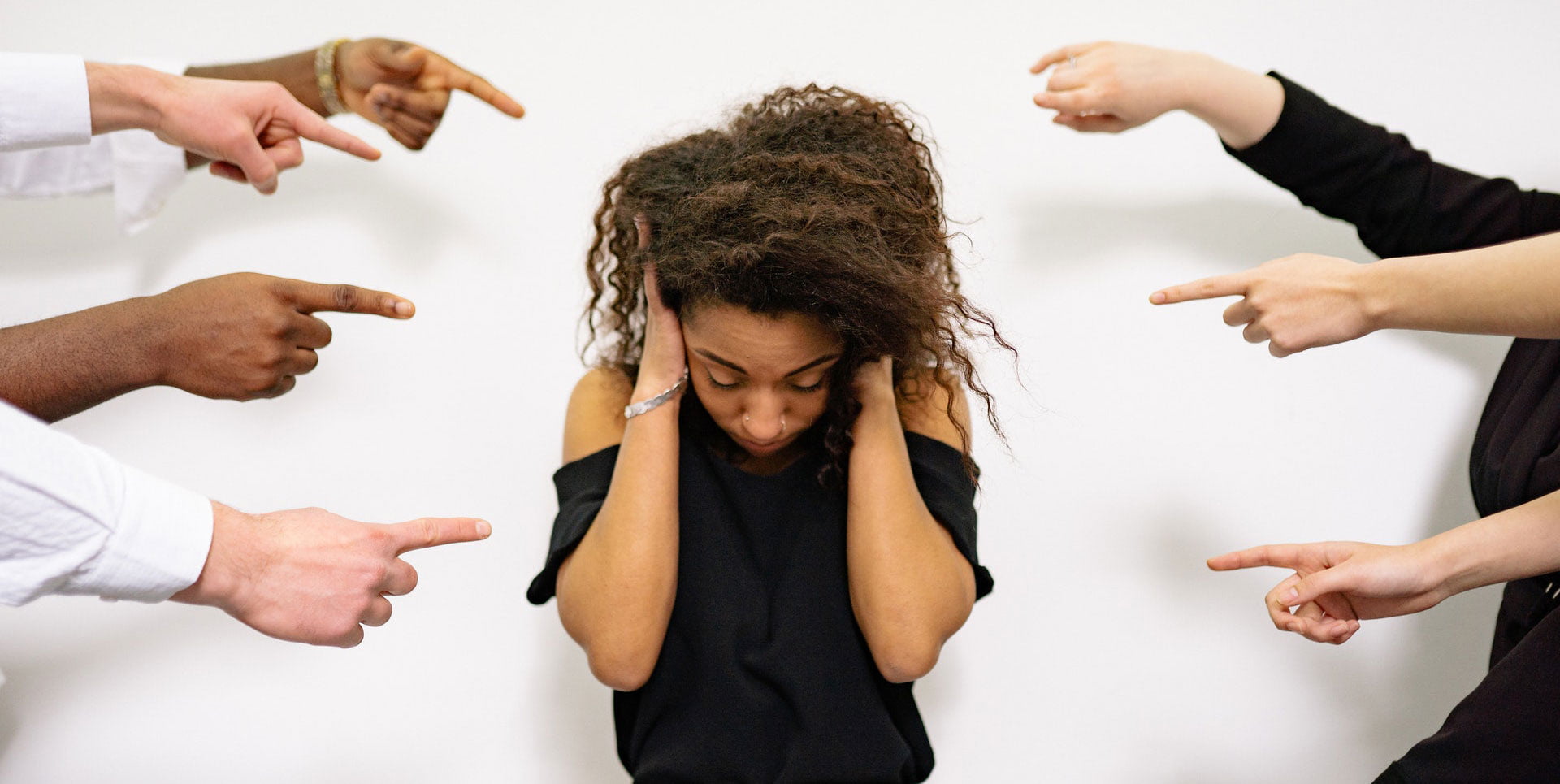 Woman covering her ears while several hands point at her, representing social pressure and anxiety.