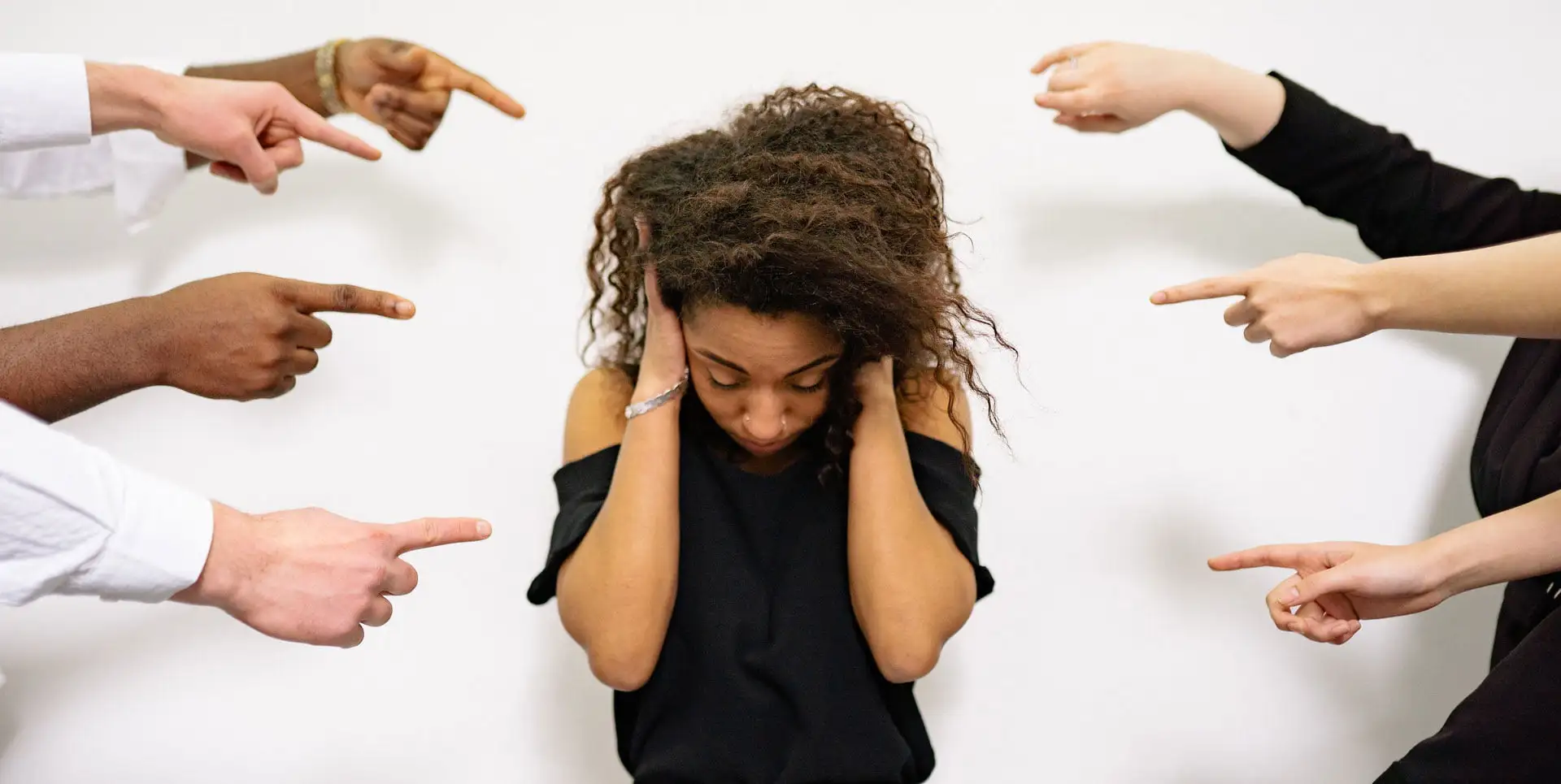 Woman covering her ears while several hands point at her, representing social pressure and anxiety.
