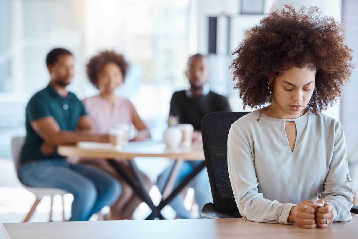 oung woman sitting alone with a sad or withdrawn expression while a blurred group of people talks in the background, conveying feelings of isolation, exclusion, or social anxiety.