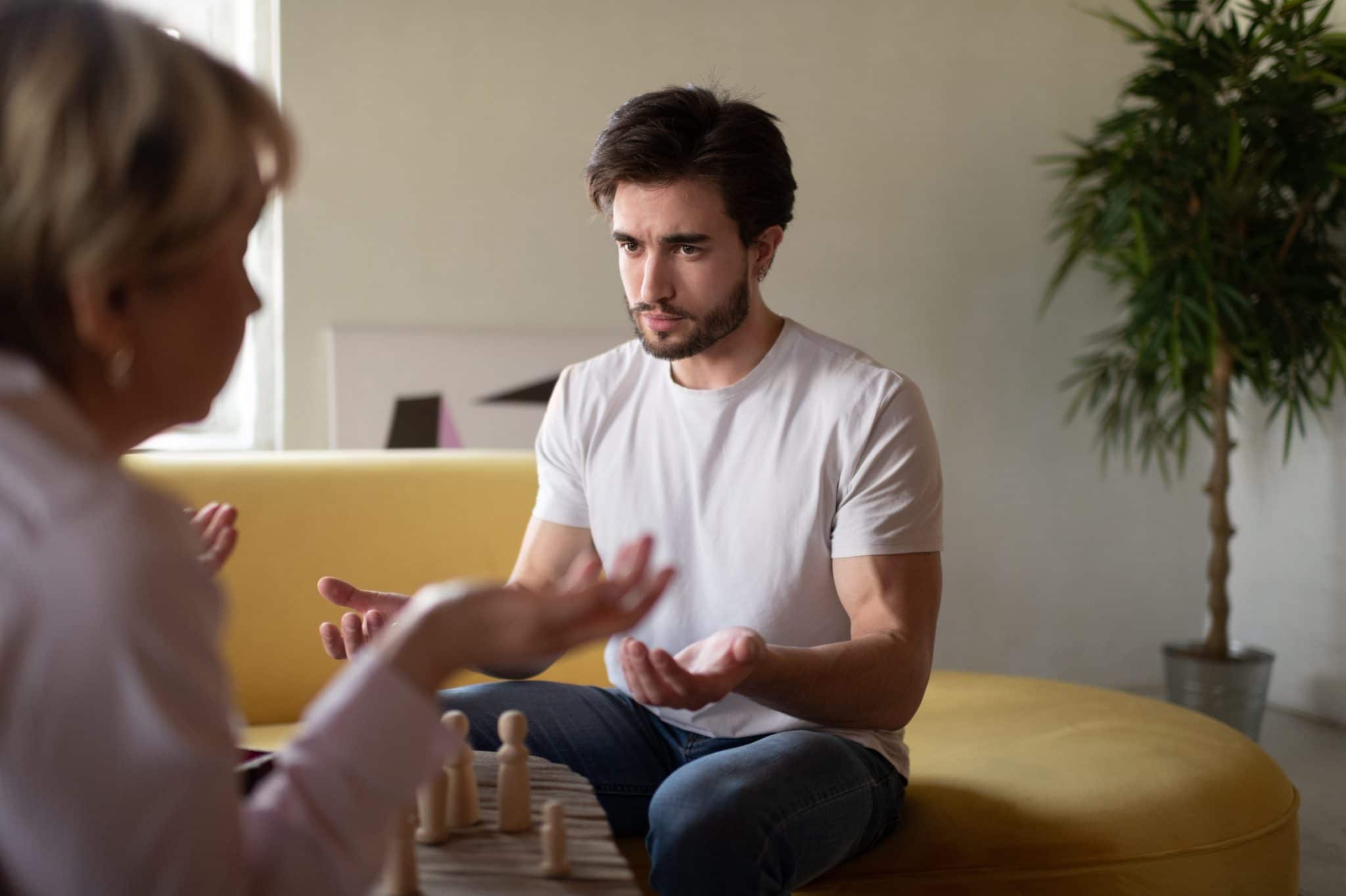 Man sitting on a yellow couch during a therapy session, expressing emotion with open hands