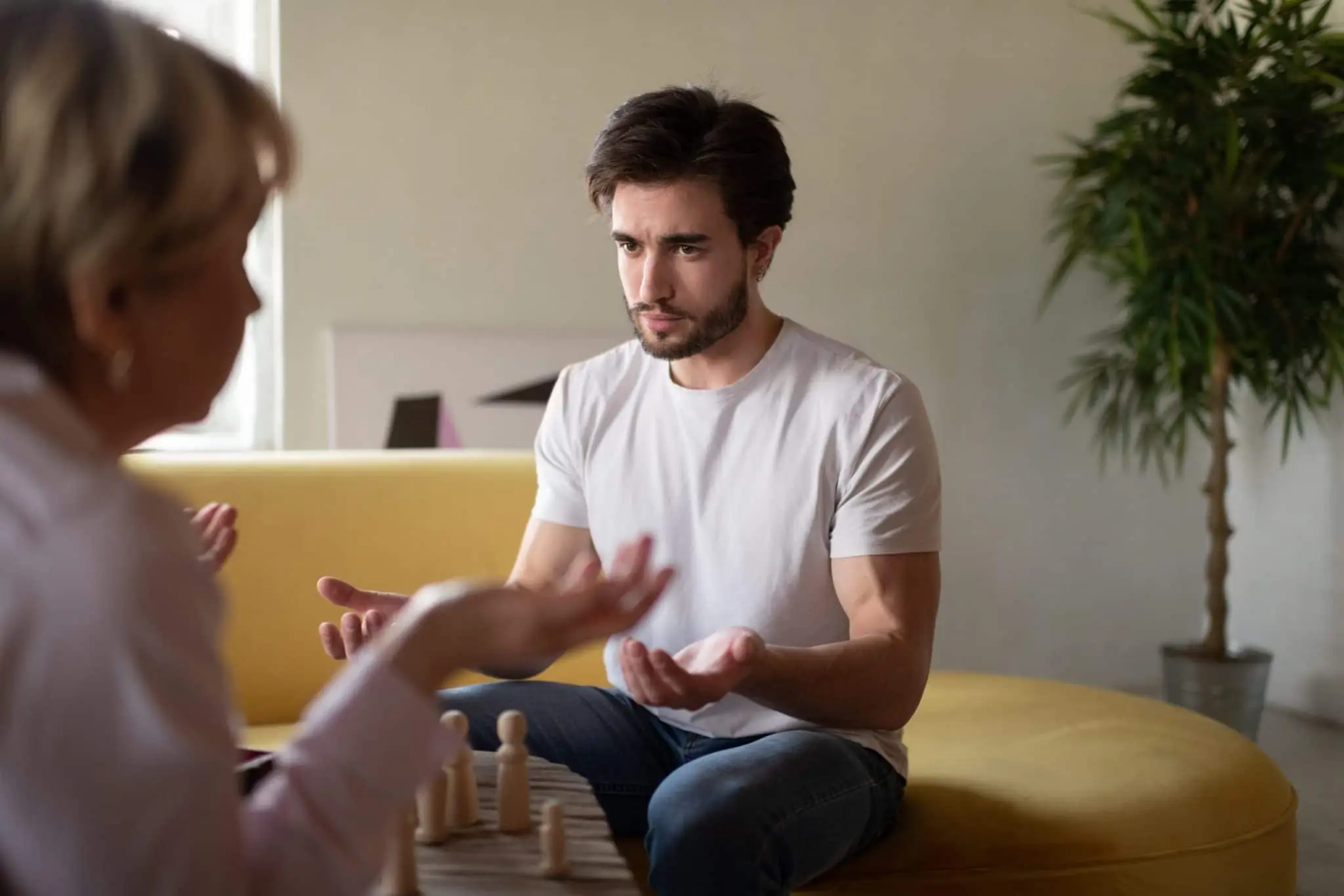 Man sitting on a yellow couch during a therapy session, expressing emotion with open hands