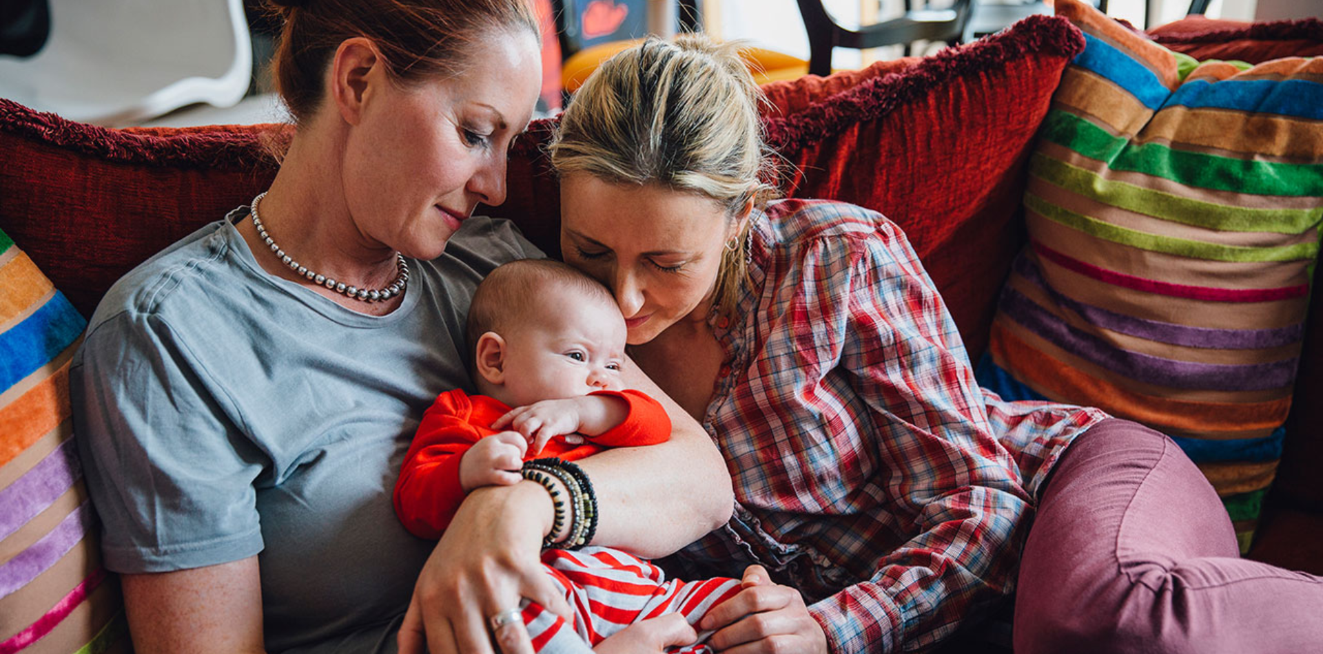 Same-sex couple lovingly holding and caring for their baby on a cozy couch with colorful pillows, highlighting their affectionate bond.