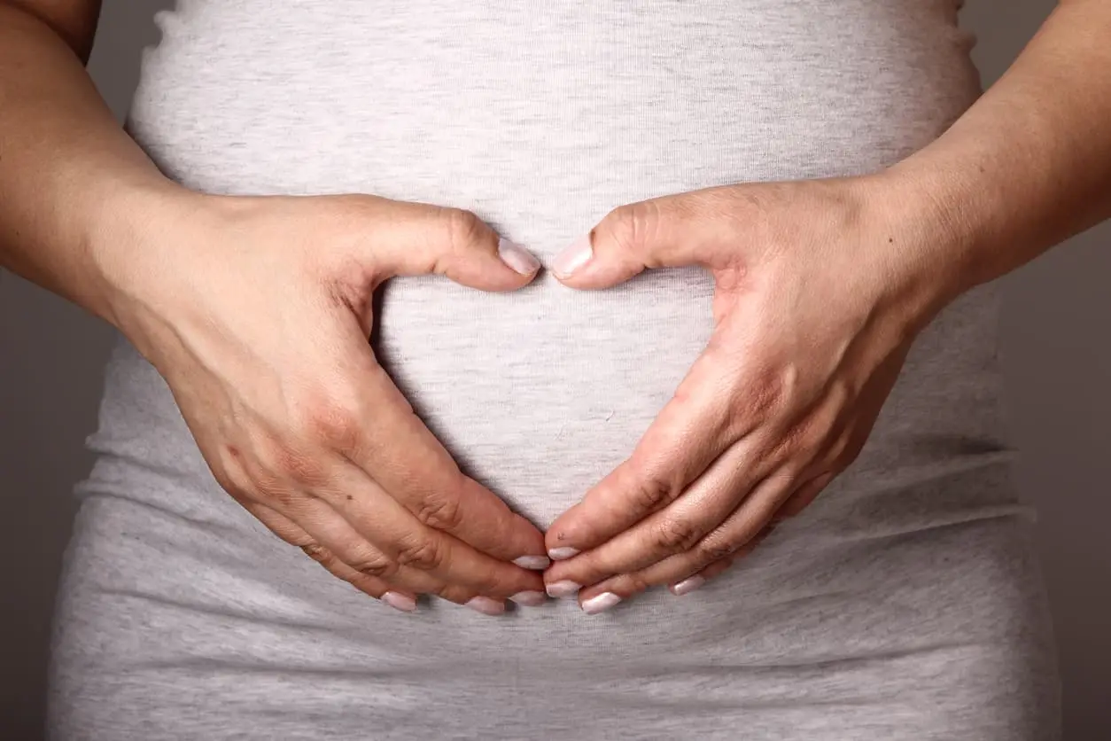 Close-up of a pregnant woman’s hands forming a heart shape over her baby bump while wearing a light gray shirt.