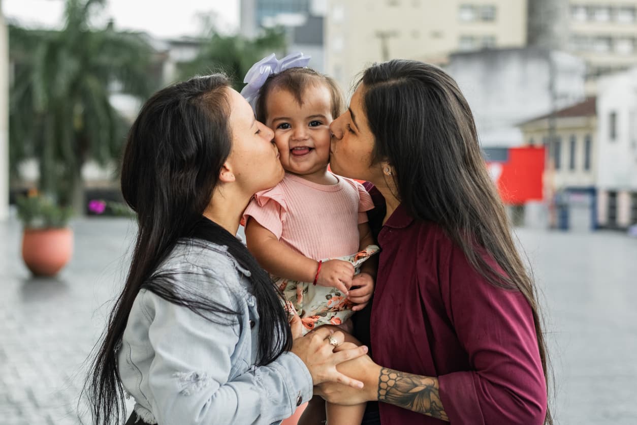 Two women affectionately kissing a smiling toddler on the cheeks while holding the child outdoors in an urban setting, capturing a joyful family moment.