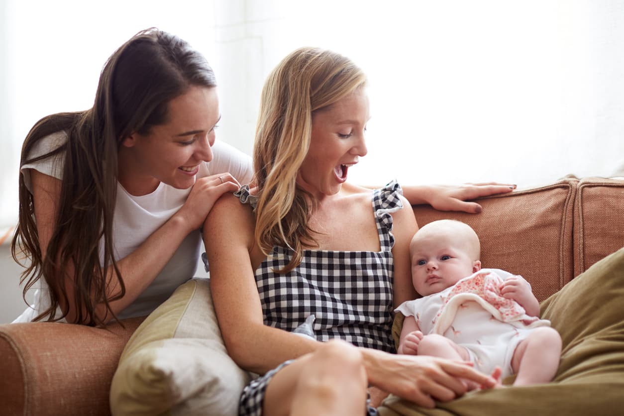 Two women sitting on a sofa smiling and interacting with a baby in a bright living room setting.