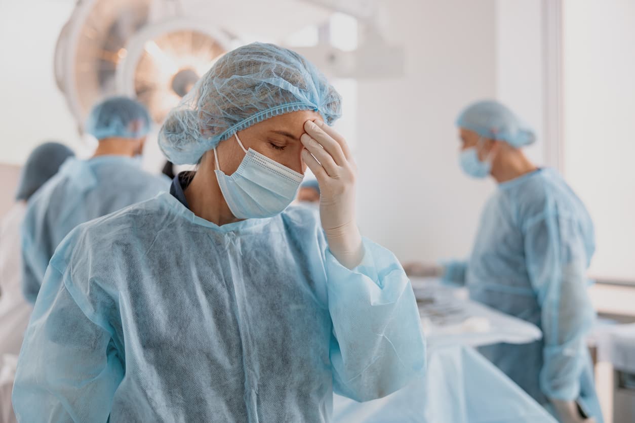 A healthcare professional in surgical protective equipment holding their forehead in apparent fatigue or stress while standing in an operating room with medical staff working in the background.