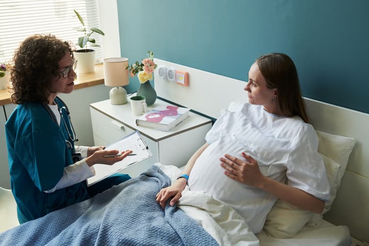 Healthcare professional speaking with a pregnant woman resting in a hospital or maternity care room, discussing prenatal care while reviewing notes on a clipboard.