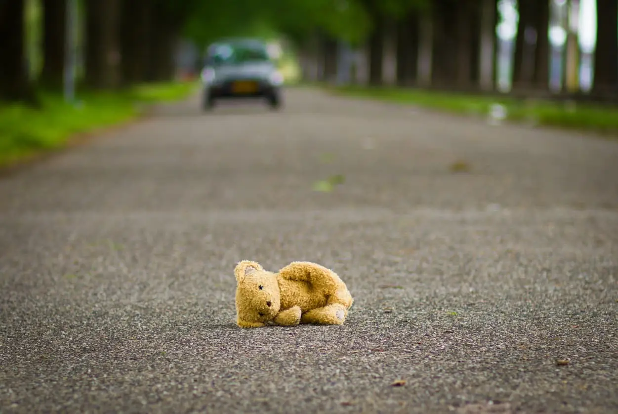Small teddy bear lying alone on an empty road with a blurred car in the distance, creating a lonely and emotional scene.