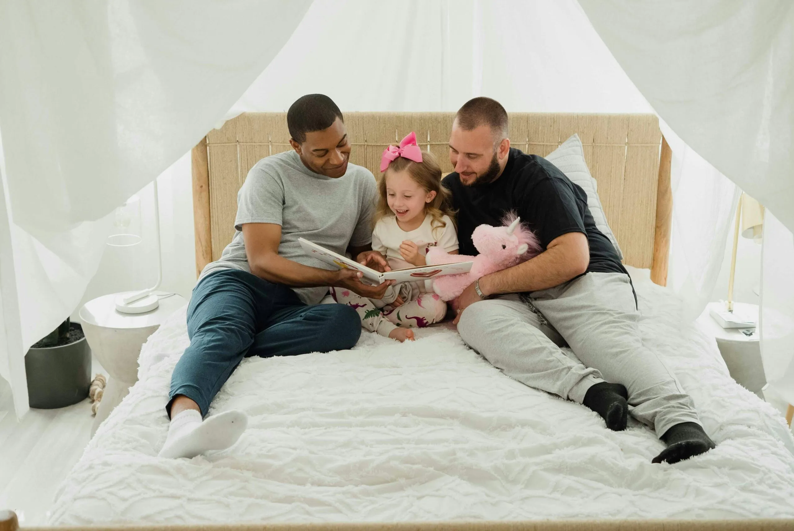 Same-sex male couple reading a book with their young daughter on a cozy bed, symbolizing love, connection, and the strength of diverse families in parenting and child development