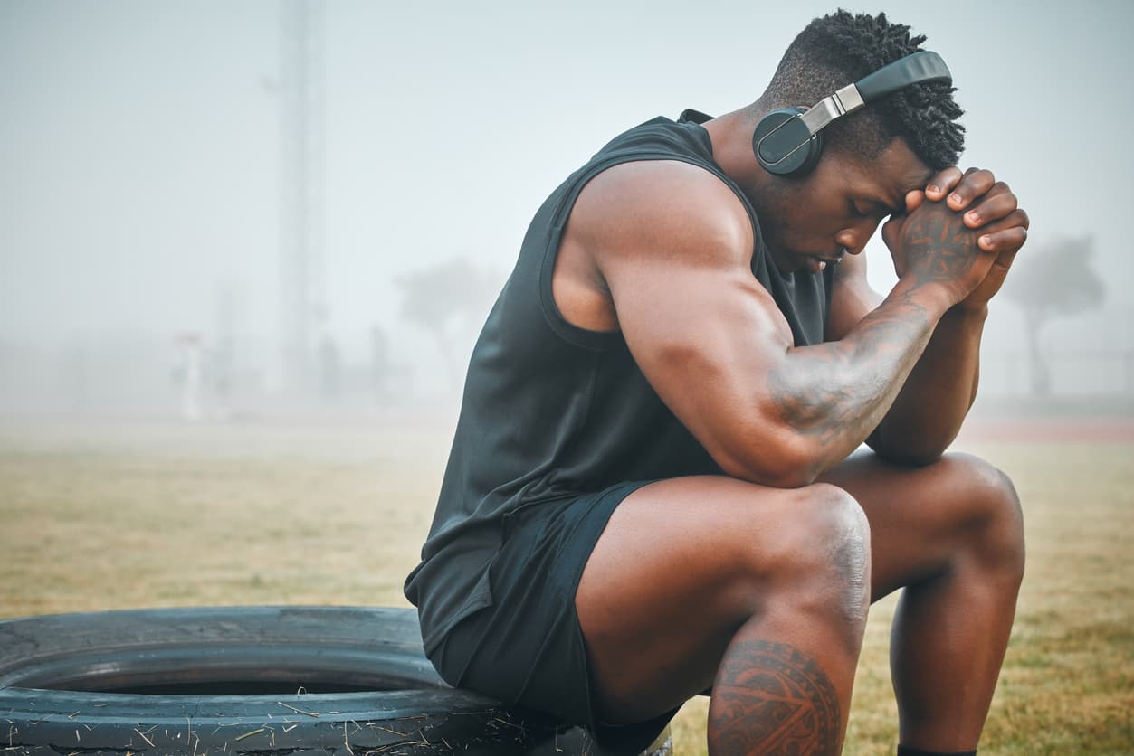 Muscular athlete wearing headphones sits on a large tire outdoors in foggy weather, leaning forward with hands clasped in a reflective or exhausted pose after exercise.