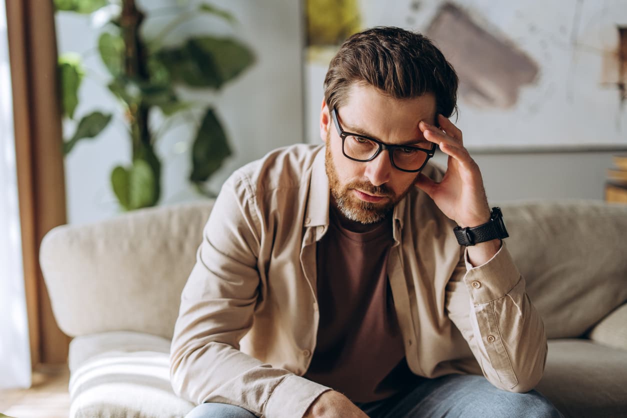 Concerned man wearing glasses sitting on a sofa with his hand on his forehead, appearing stressed, worried, or deep in thought in a home setting.