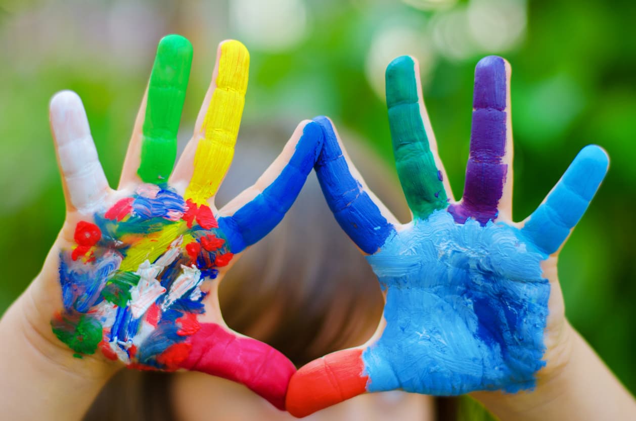 Close-up of a child’s hands covered in bright, colorful paint, forming the “I love you” sign against a soft green outdoor background, symbolizing creativity, self-expression, inclusion, and communication.