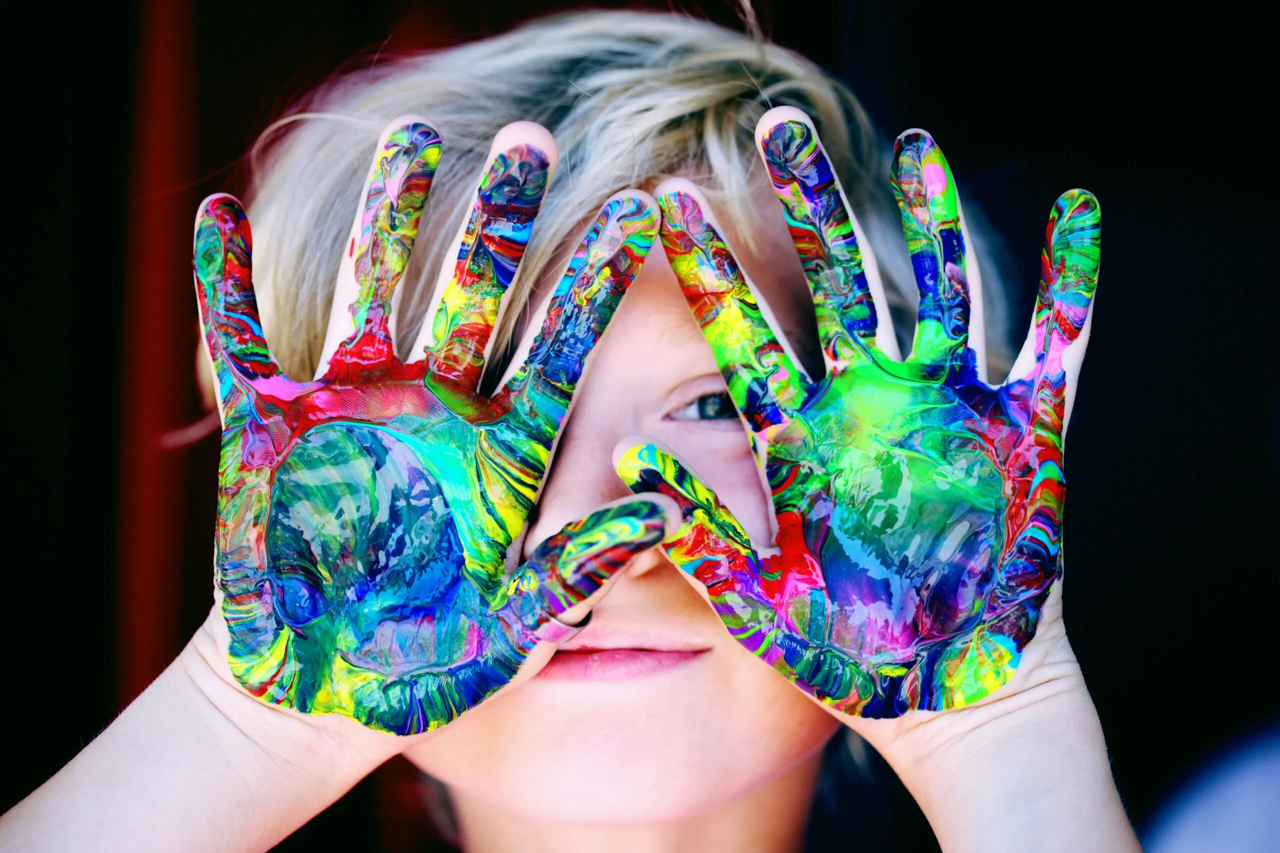 A child with brightly painted hands held up to their face, smiling through the colors.