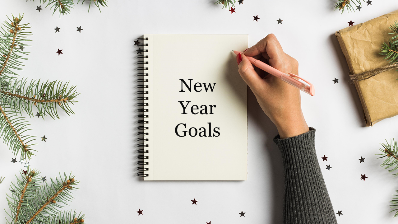 A hand writing in a notebook labeled "New Year Goals" surrounded by pine branches and stars.