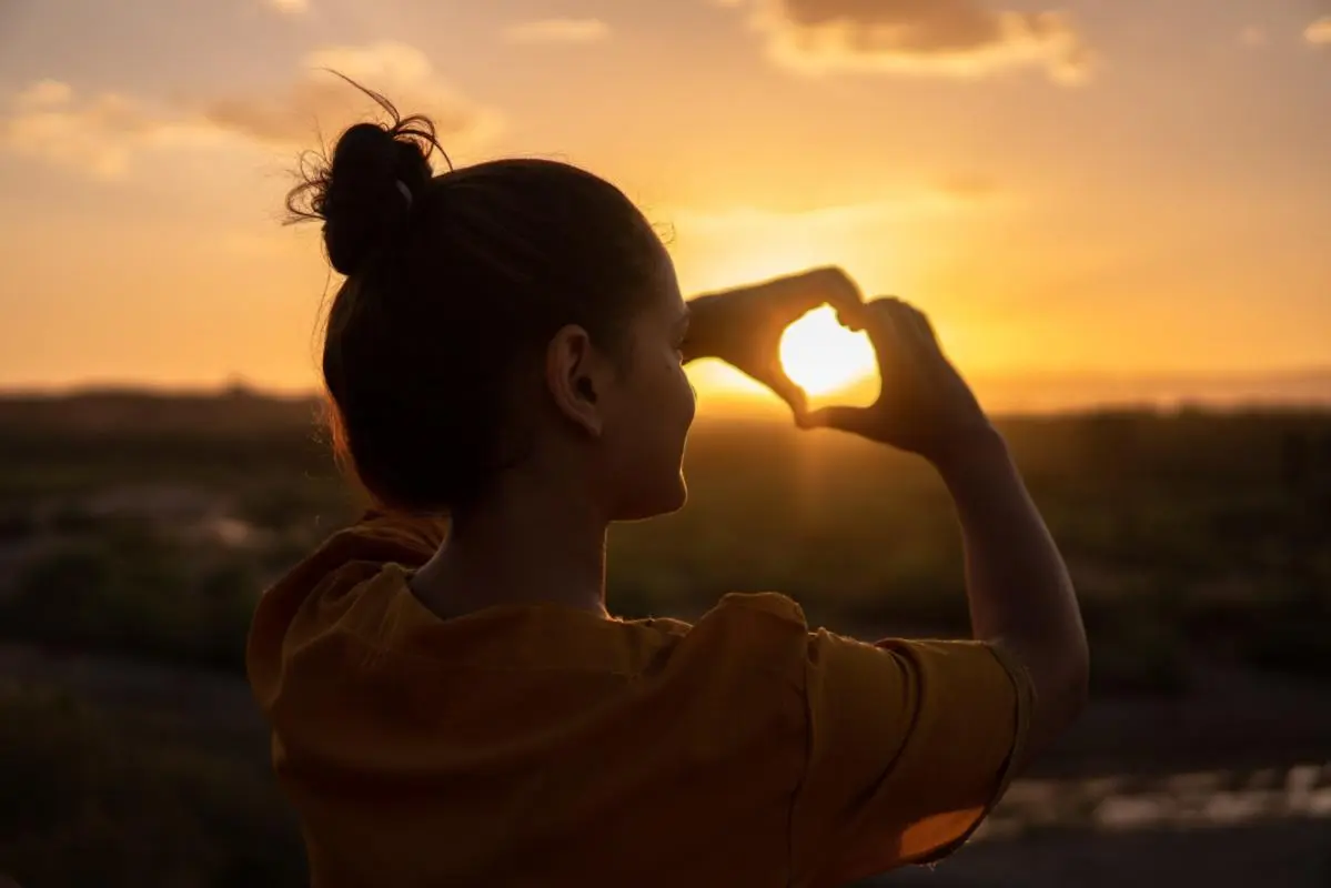 Woman forming a heart shape with her hands at sunset, symbolizing the power of self-love, inner peace, and emotional healing through self-compassion