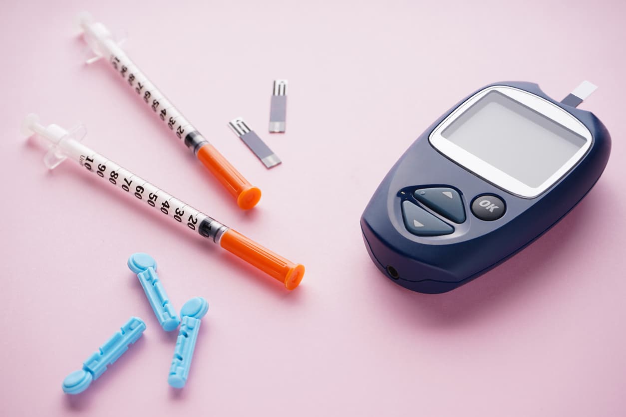 Blood glucose meter with test strips, lancets, and insulin syringes arranged on a pink background, representing diabetes testing and management supplies.