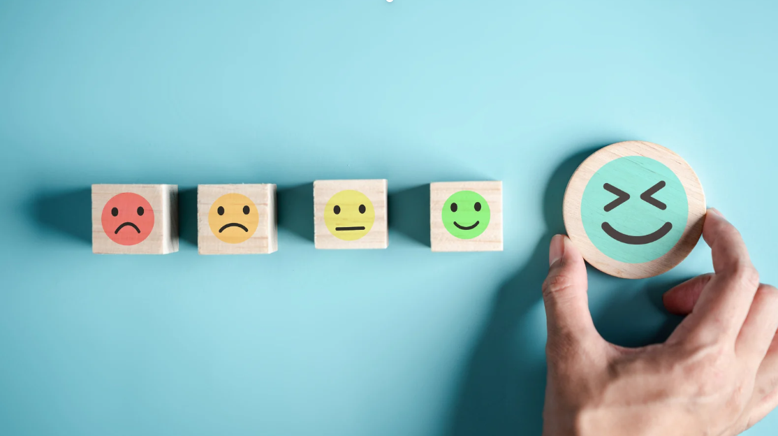Wooden blocks displaying emoji faces ranging from sad to happy on a blue background, with a hand placing a laughing face at the end of the row representing emotional progress and mental wellness.