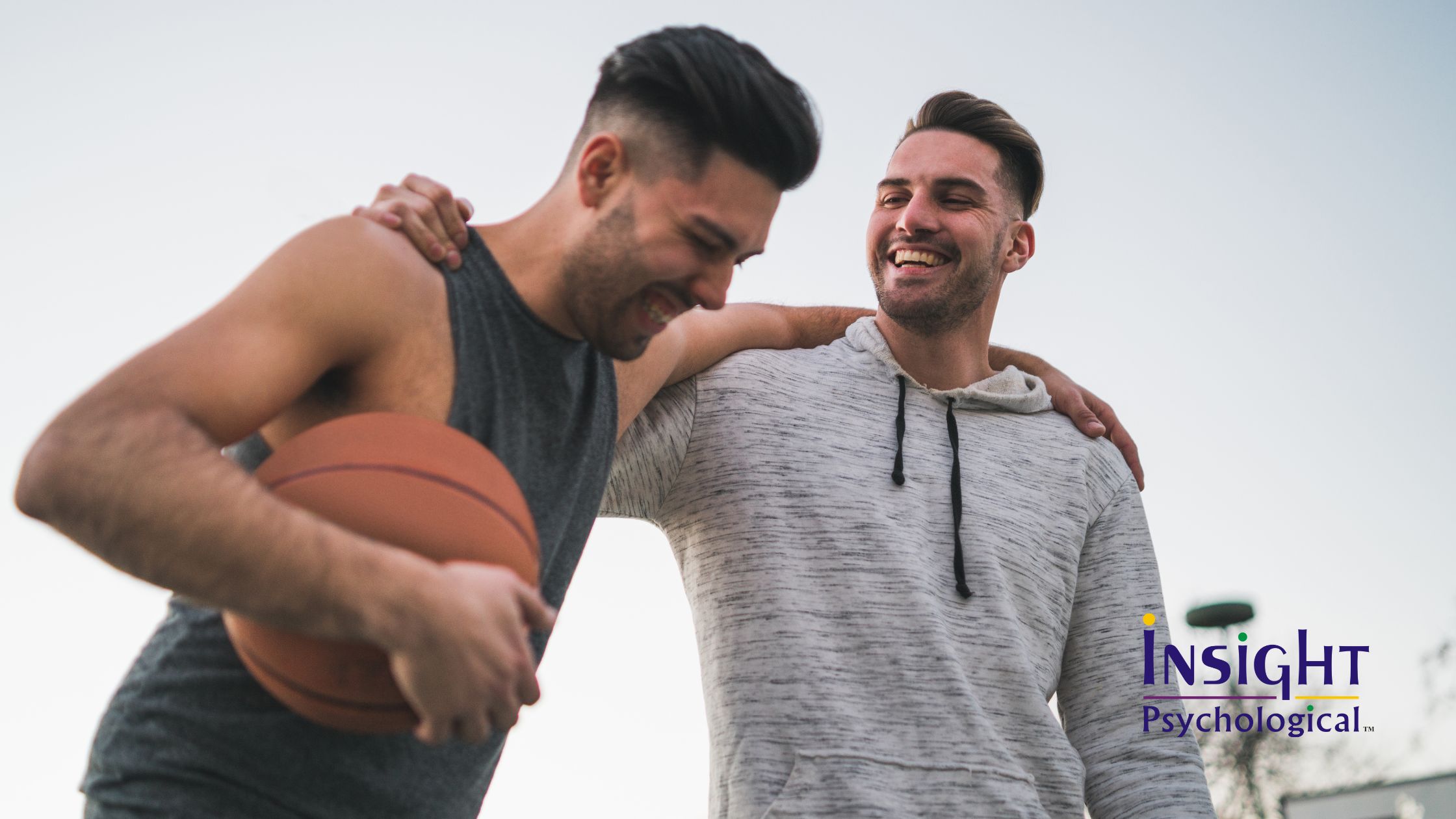 Two men laughing together on a basketball court, one holding a basketball and the other with his arm around his friend's shoulder, representing male friendship and connection in support of men's mental health.